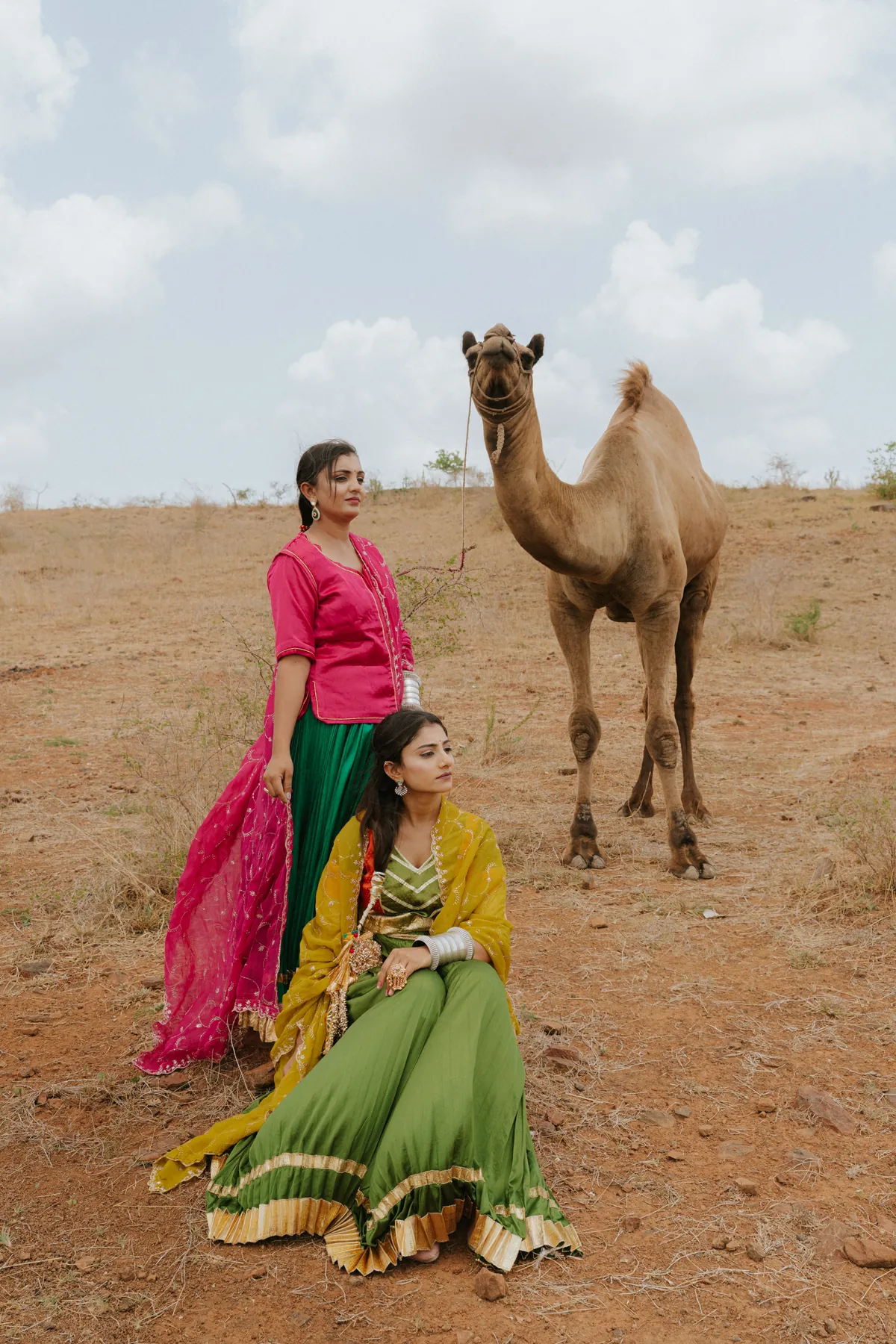 Two women in colourful traditional outfits posing beside a camel