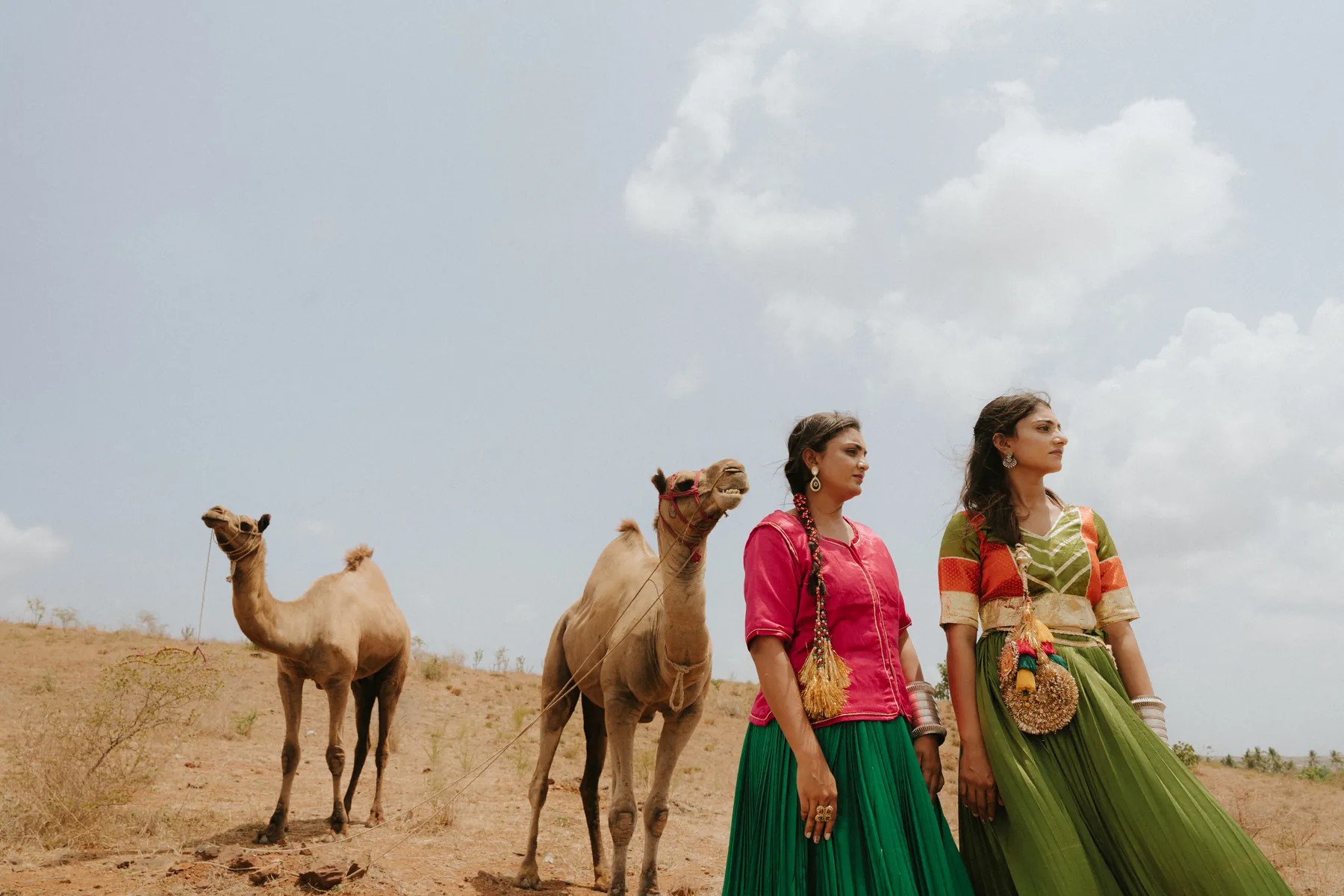 Two women in colourful traditional outfits posing with camels in an outdoor fashion shoot